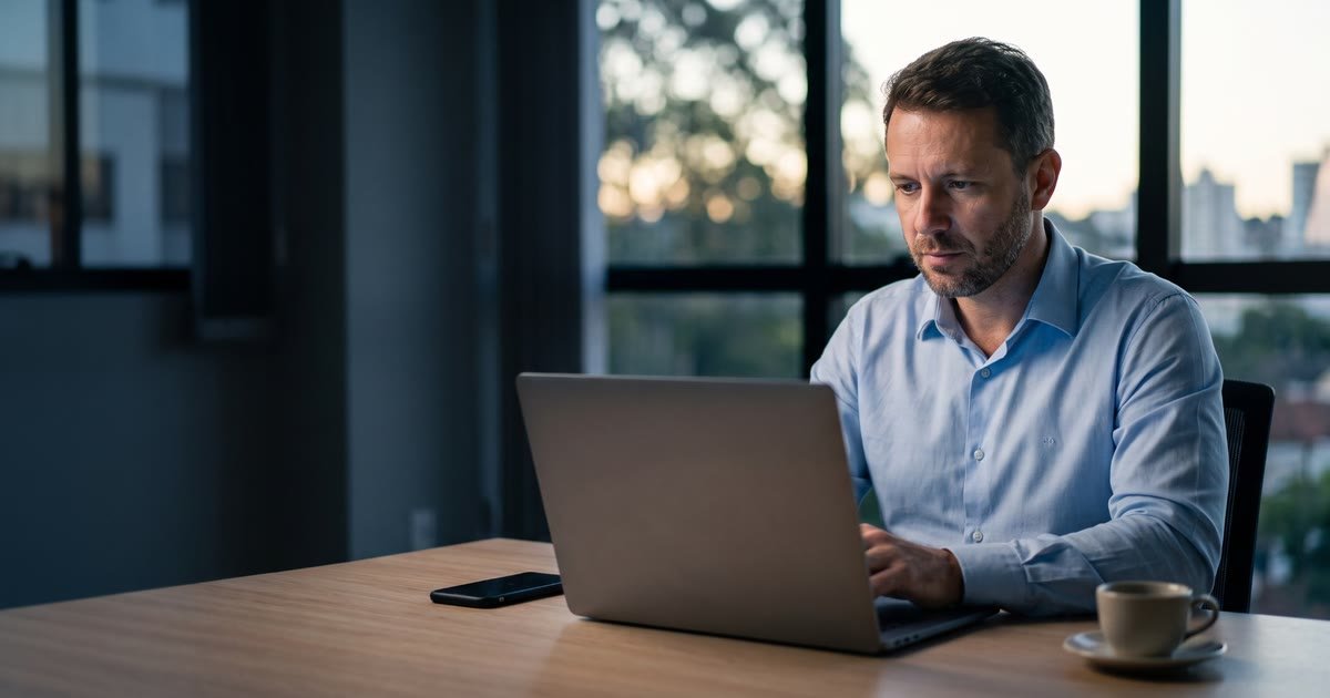 Empresário de camisa azul-clara trabalhando no notebook em um escritório no fim da tarde, com a cidade ao fundo pela janela